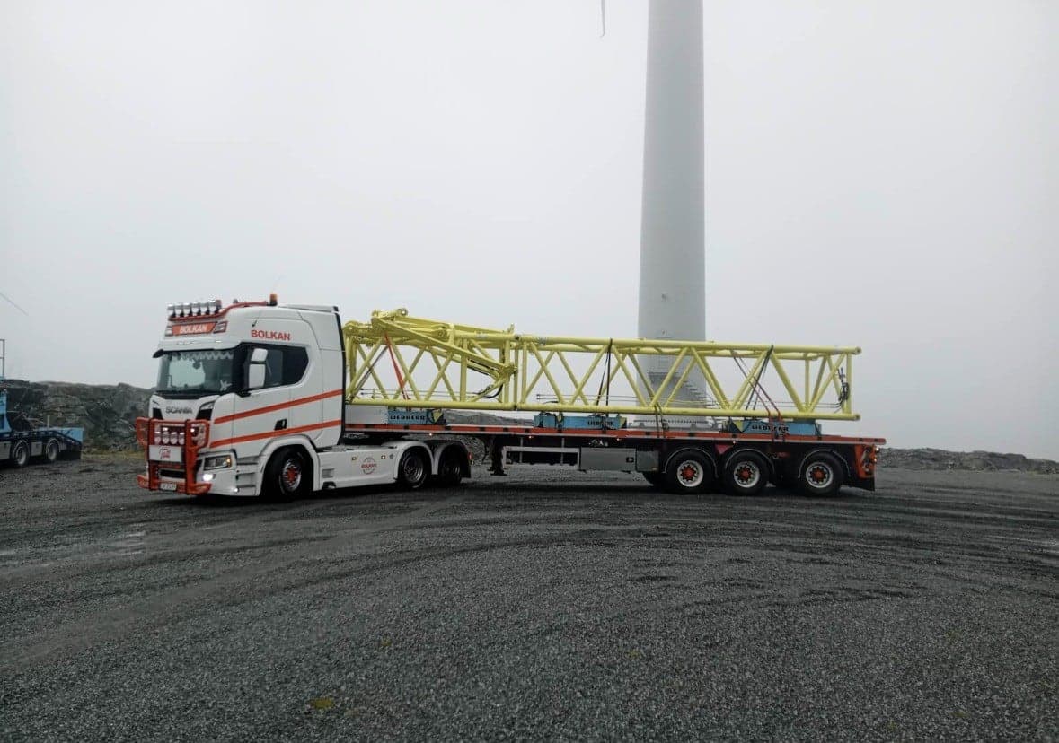 Hvit Bolkan Scania med gul kranbom på hengeren ved en vindmølle