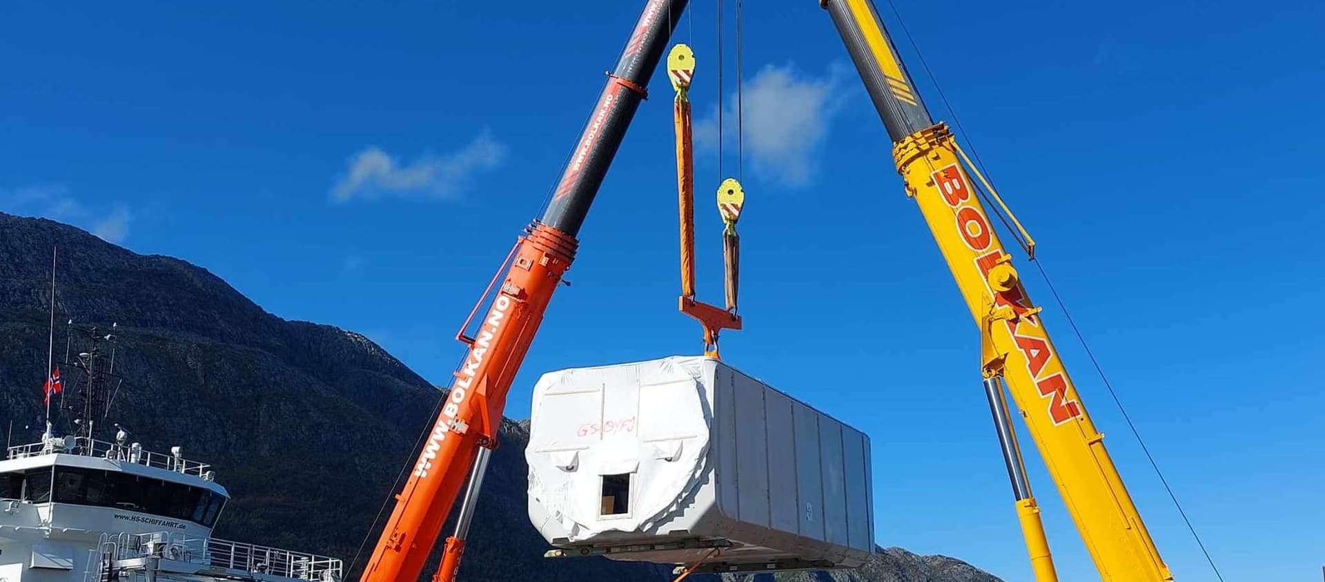 To Bolkan mobilkraner løfter sammen en stor modul fra et skip ved kaia, under blå himmel