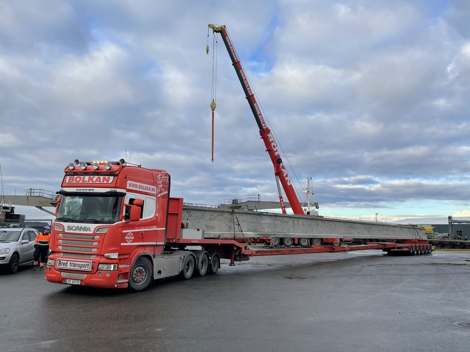 Bolkan Scania med skiltet Bred transport og en lang betongbjelke på hengeren, mens en mobilkran løfter i bakgrunnen