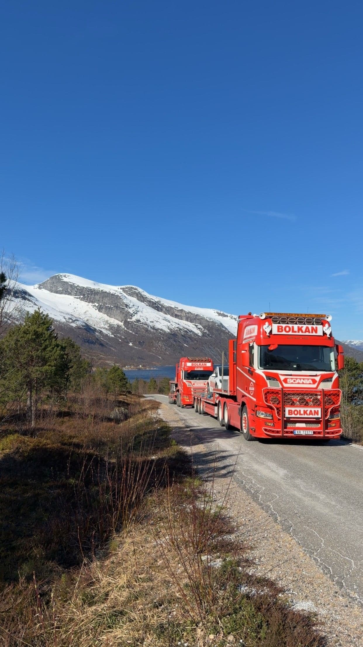 To Bolkan Scania-vogntog i kolonne ved et snødekt fjell og en fjord