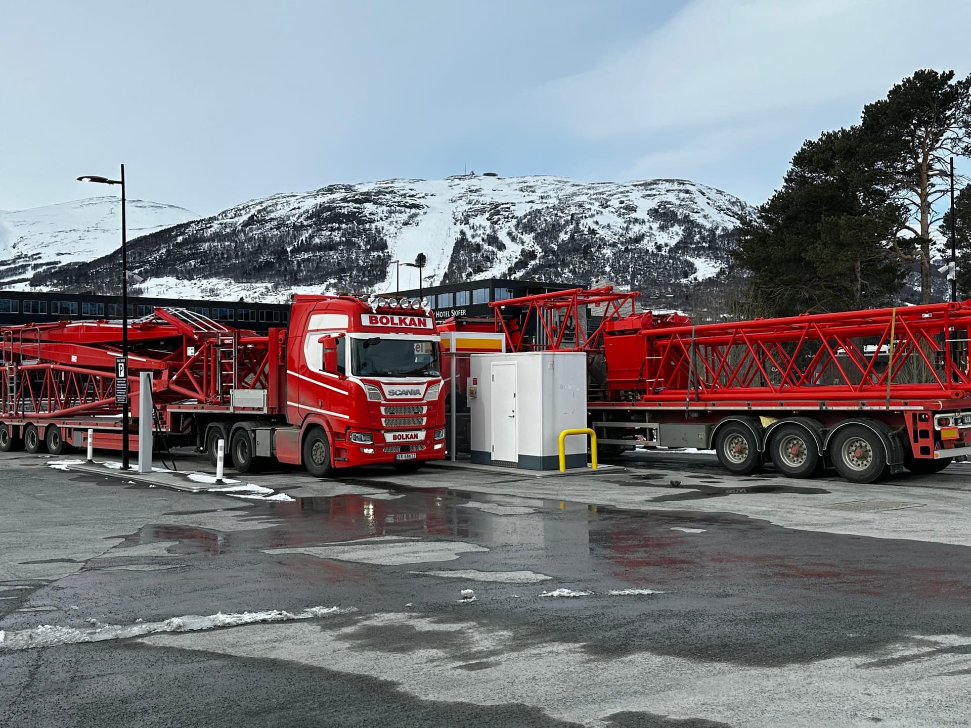 To Bolkan-semitrailere lastet med røde kranbomdeler ved en bensinstasjon, med snødekte fjell bak